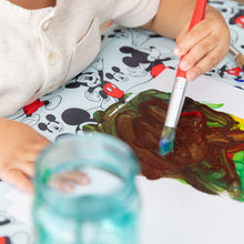 Load image into Gallery viewer, Close-up of a child painting with brown and green paint on paper on Bumkins Splat Mat: Mickey Mouse.
