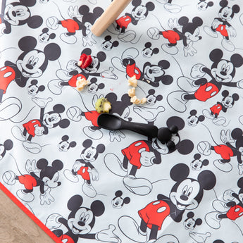 A black spoon and scattered food sits on top of Bumkins Splat Mat: Mickey Mouse on a floor under a chair.