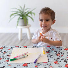 Load image into Gallery viewer, A smiling child with short brown hair sits at a table covered by Bumkins Splat Mat: Mickey Mouse, coloring with markers on paper.
