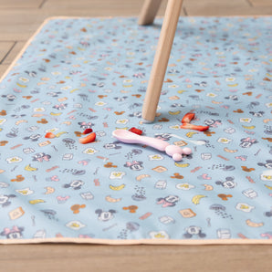 Bumkins Splat Mat: Mickey + Minnie Sunny Side Up lays on a wood floor under a wooden chair leg, with strawberry pieces and a spoon on top.