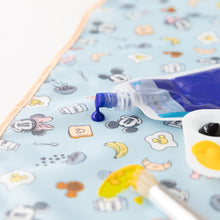 Load image into Gallery viewer, A blue paint tube spilled on a Bumkins Splat Mat: Mickey + Minnie Sunny Side Up, near a palette with yellow and black paint and a brush.
