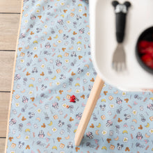 Load image into Gallery viewer, Bumkins Splat Mat: Mickey + Minnie Sunny Side Up lays on a wood floor underneath a high chair and strawberry pieces.
