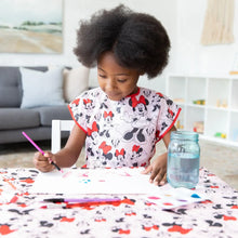 Load image into Gallery viewer, A child paints at a table covered by Bumkins Splat Mat: Minnie Mouse, wearing a matching smock, in a cozy living room.
