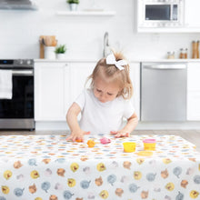 Load image into Gallery viewer, A toddler with a white bow plays with colorful clay on a Bumkins Splat Mat: Winnie and Friends in a bright kitchen.
