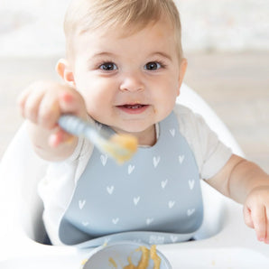 A smiling baby wearing a Bumkins Silicone Bib: Hearts in a high chair smiles, holding a gray spoon with applesauce to the camera.