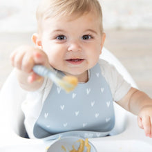 Load image into Gallery viewer, A smiling baby wearing a Bumkins Silicone Bib: Hearts in a high chair smiles, holding a gray spoon with applesauce to the camera.
