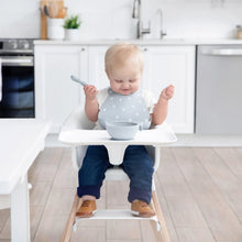 Load image into Gallery viewer, A baby sits in a high chair wearing a Bumkins Silicone Bib: Hearts, looking down at a gray bowl as they smile in a bright kitchen.

