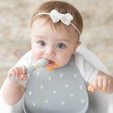 Load image into Gallery viewer, A baby with a white bow and a messy face in a high chair wears a Bumkins Silicone Bib: Hearts while chewing on a gray spoon.
