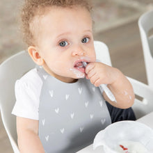 Load image into Gallery viewer, A curly-haired toddler with a messy face eats with a spoon, wearing a Bumkins Silicone Bib: Hearts.

