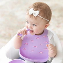 Load image into Gallery viewer, A baby in a bow headband chews a purple spoon, wearing a Bumkins Silicone Bib: Rainbows while seated in a high chair.
