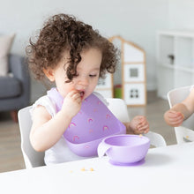 Load image into Gallery viewer, A toddler with curly hair wearing a Bumkins Silicone Bib: Rainbows enjoys a meal from a purple bowl at a white table in a bright playroom.
