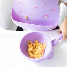 Load image into Gallery viewer, Close-up of a child in a Bumkins Silicone Bib: Rainbows enjoying mac and cheese from a matching purple bowl on a high chair tray.
