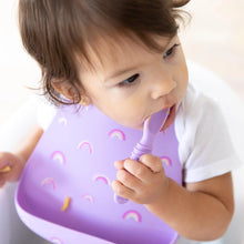 Load image into Gallery viewer, Close-up of a toddler wearing a Bumkins Silicone Bib: Rainbows while chewing a purple spoon.
