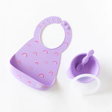 Load image into Gallery viewer, Bumkins Silicone Bib: Rainbows lays next to a purple bowl &amp; spoon set on a white background.
