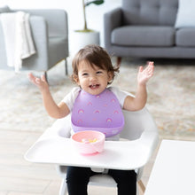 Load image into Gallery viewer, A toddler in a high chair, wearing a Bumkins Silicone Bib: Rainbows, smiles with raised arms with a pink bowl nearby. 
