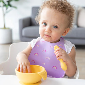 A curly-haired baby in a Bumkins Silicone Bib: Rainbows holds a spoon, ready to eat from a yellow bowl, with a couch in the background.