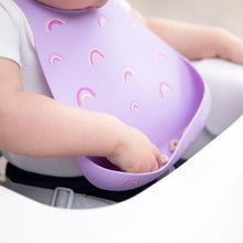 Load image into Gallery viewer, A baby in a high chair wears a Bumkins Silicone Bib: Rainbows, reaching for cereal in the pocket.
