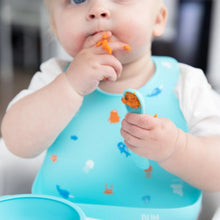 Load image into Gallery viewer, A baby wearing a Bumkins Silicone Bib: Ocean Life eats mashed sweet potato with their hands and a blue spoon.
