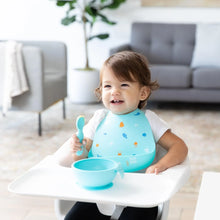 Load image into Gallery viewer, A toddler in a high chair smiles in a bright living room, wearing a Bumkins Silicone Bib: Ocean Life and holding a blue spoon near a bowl.
