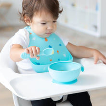Load image into Gallery viewer, A toddler in a Bumkins Silicone Bib: Ocean Life holds a blue spoon and eyes a blue bowl of food on a high chair tray.
