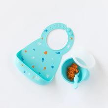 Load image into Gallery viewer, Bumkins Silicone Bib: Ocean Life lays next to a blue bowl &amp; spoon set with orange mashed food on a white background.

