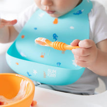 Load image into Gallery viewer, Close-up of a baby in a high chair wearing a Bumkins Silicone Bib: Ocean Life, eating yogurt from an orange bowl with a spoon.
