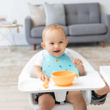 Load image into Gallery viewer, A baby in a high chair smiles during mealtime, wearing a Bumkins Silicone Bib: Ocean Life and eating from an orange bowl.
