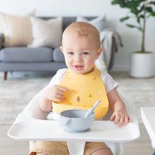 Load image into Gallery viewer, A smiling toddler in a high chair wears a Bumkins Silicone Bib: Camp Gear, with a gray bowl and spoon in front of them on the tray.
