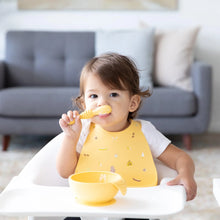 Load image into Gallery viewer, A toddler sits in a high chair wearing a Bumkins Silicone Bib: Camp Gear, enjoying baby-led weaning from a yellow bowl with a spoon.
