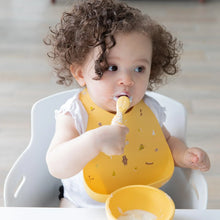 Load image into Gallery viewer, A curly-haired toddler with a messy face in a high chair wears a Bumkins Silicone Bib: Camp Gear, eating yogurt from a yellow bowl.
