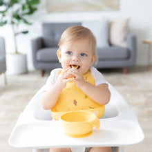 Load image into Gallery viewer, A smiling toddler sits in a high chair wearing a Bumkins Silicone Bib: Camp Gear, exploring self-feeding from a yellow bowl with a spoon.
