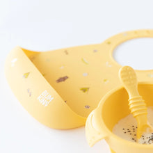 Load image into Gallery viewer, Close-up of Bumkins Silicone Bib: Camp Gear a yellow bowl with yogurt, and a yellow spoon on a white background.
