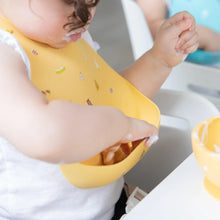 Load image into Gallery viewer, A toddler in a Bumkins Silicone Bib: Camp Gear messily enjoys creamy food as they reach into the front pocket of the bib.

