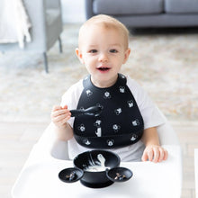 Load image into Gallery viewer, A smiling toddler in a high chair wears a Bumkins Silicone Bib: Mickey Mouse Faces, holding a spoon by a food-filled bowl on the tray.
