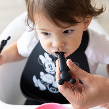 Load image into Gallery viewer, Close-up of a hand feeding a child wearing a black bib in a high chair with a Bumkins Silicone Dipping Spoons: Minnie Mouse (Black and Pink).
