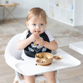 A toddler in a high chair, wearing a Bumkins Silicone Bib: Mickey + Minnie Mouse, eagerly eats from a gold bowl with a gold spoon.
