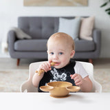 A baby in a high chair wears a Bumkins Silicone Bib: Mickey + Minnie Mouse, holding a spoon near a gold bowl; a gray sofa in the background.