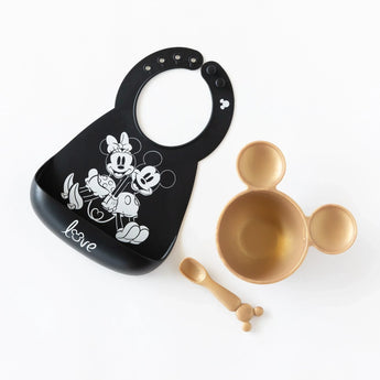 Bumkins Silicone Bib: Mickey + Minnie Mouse lays on a white background next to a gold Mickey-shaped bowl and matching spoon.