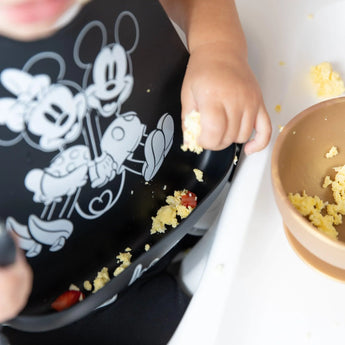Close-up of a child wearing a Bumkins Silicone Bib: Mickey + Minnie Mouse, with focus on scrambled eggs in the front pocket.