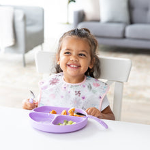 Load image into Gallery viewer, A smiling girl in a floral bib uses Bumkins Spoon + Fork: Lavender to eat from a matching plate at a white table.
