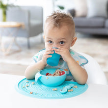 Load image into Gallery viewer, A toddler sips from a Bumkins Silicone Starter Cup: Blue at a white table, wearing a patterned bib with a matching bowl and food mat.
