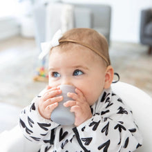 Load image into Gallery viewer, A baby wearing a white &amp; black bib and a bow in a high chair sips from a Bumkins Silicone Starter Cup: Gray.
