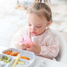 Load image into Gallery viewer, A toddler in a pink bib holds a Bumkins Silicone Starter Cup: Pink, sitting at a table with a gray plate of fruits and snacks.
