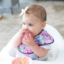 Load image into Gallery viewer, A baby in a bib and bow headband sits in a high chair, holding a Bumkins Silicone Starter Cup: Pink beside snacks.
