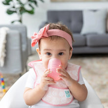 Load image into Gallery viewer, A baby in a pink headband and bib drinks from a Bumkins Silicone Starter Cup: Pink while sitting in a high chair. A gray sofa is behind.
