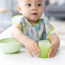 Load image into Gallery viewer, A baby wearing a cactus-print bib in a high chair reaches for a Bumkins Silicone Starter Cup: Sage next to a green bowl.
