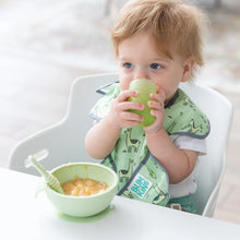 Load image into Gallery viewer, A toddler with brown hair in a high chair drinks from a Bumkins Silicone Starter Cup: Sage with a green bowl of applesauce nearby.
