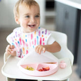 A toddler with a colorful bib in a high chair lifts a Bumkins Silicone Stretch Lid for Grip Dish 3 Section, Grip Plate from a pink plate.