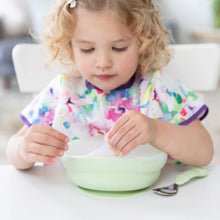 Load image into Gallery viewer, A curly-haired child in a colorful bib sits at a white table, removing a Bumkins Silicone Stretch Lid for Grip Bowl from a green bowl.
