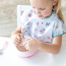 Load image into Gallery viewer, A young girl in a floral bib removes a Bumkins Silicone Stretch Lid for Grip Bowl  from a pink bowl of cereal and milk at a white table.
